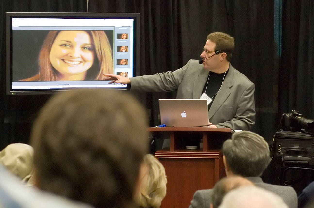 Scott Kelby speaking at a conference lectern wearing a headset microphone and pointing at a large screen showing a close-up portrait photo