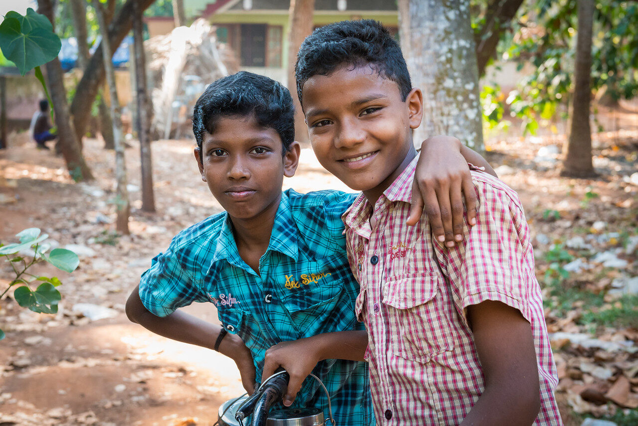 Two smiling boys with their arms around each other, in a teal checked shirt and a pink checked shirt, outdoors in India