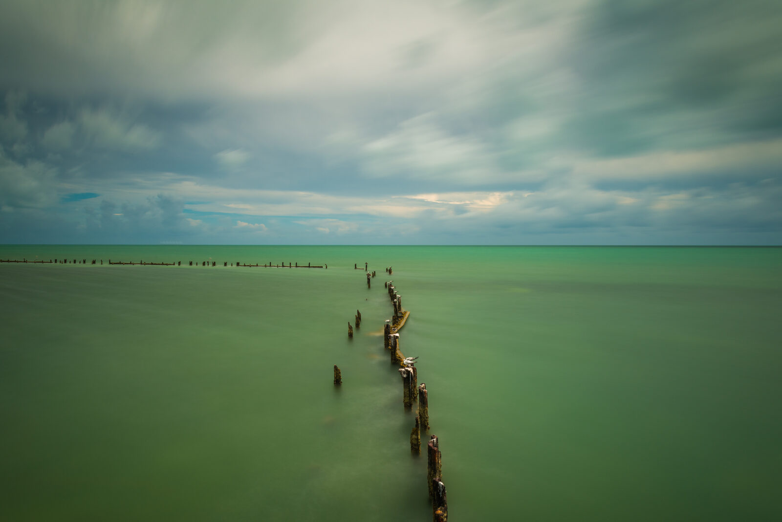 Edin Chavez long exposure of old pilings in green water beneath moving clouds