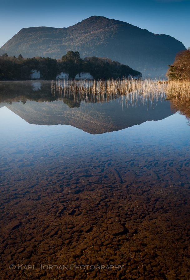 Clear shallow water over brown rocks with mountain reflection at Lough Leane, Killarney
