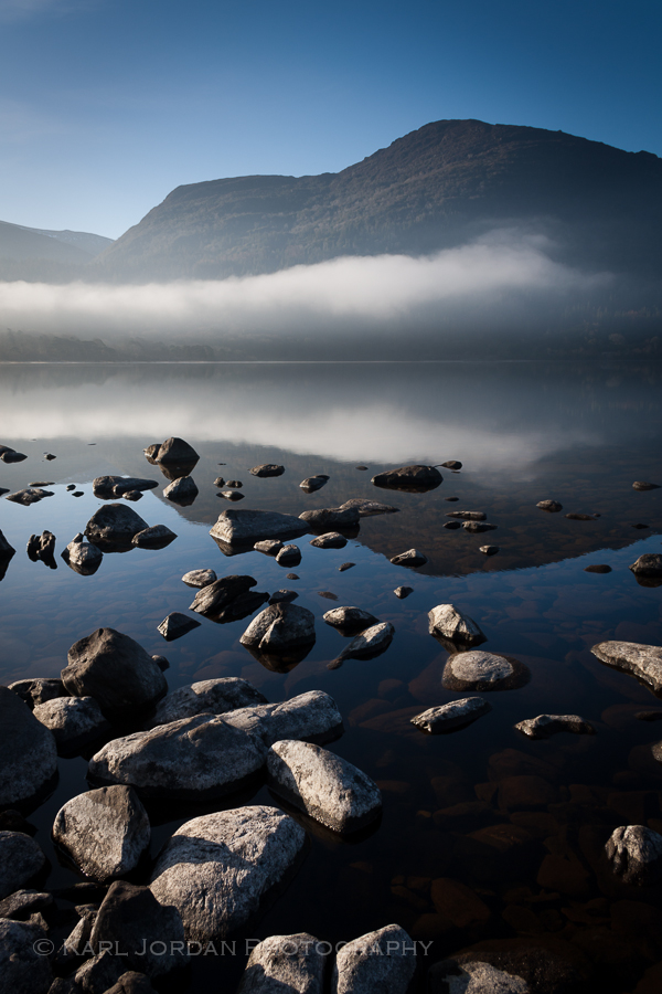 Low mist over the mountain above dark rocks in still water, Lough Leane Ireland