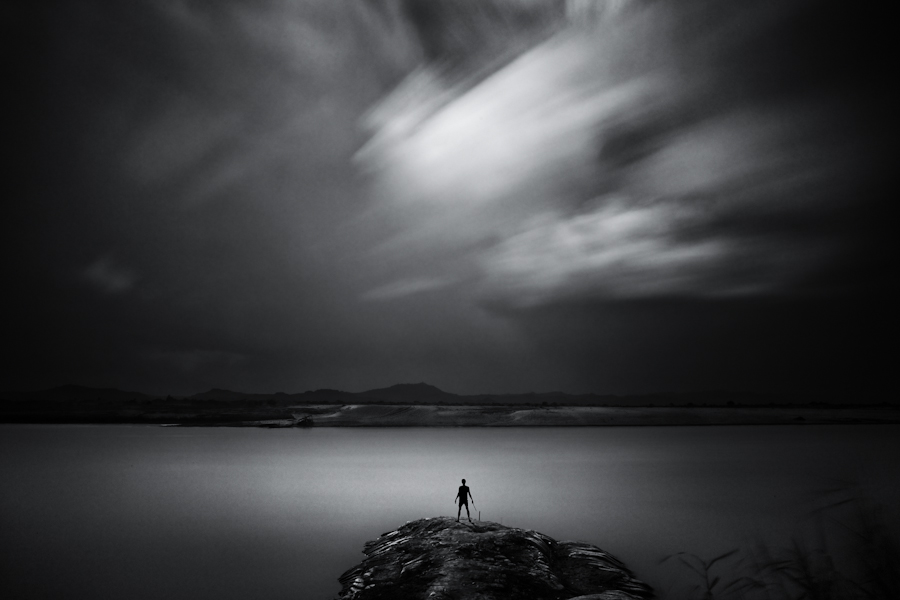 Lone figure on rock beneath streaked clouds