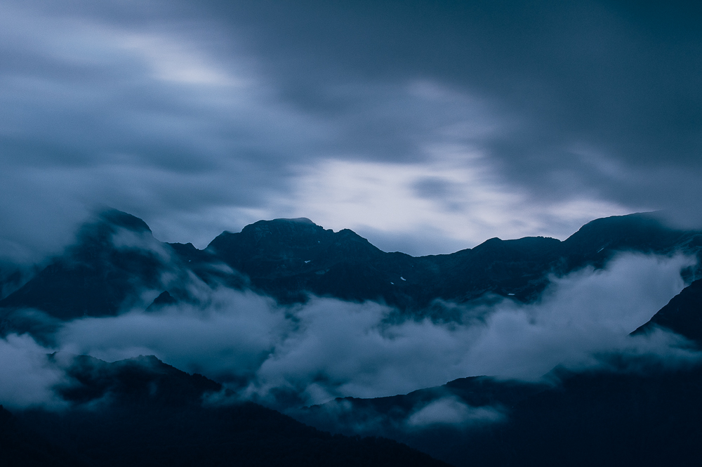 Cloud and fog moving across a dark mountain range beneath a pale blue-gray sky