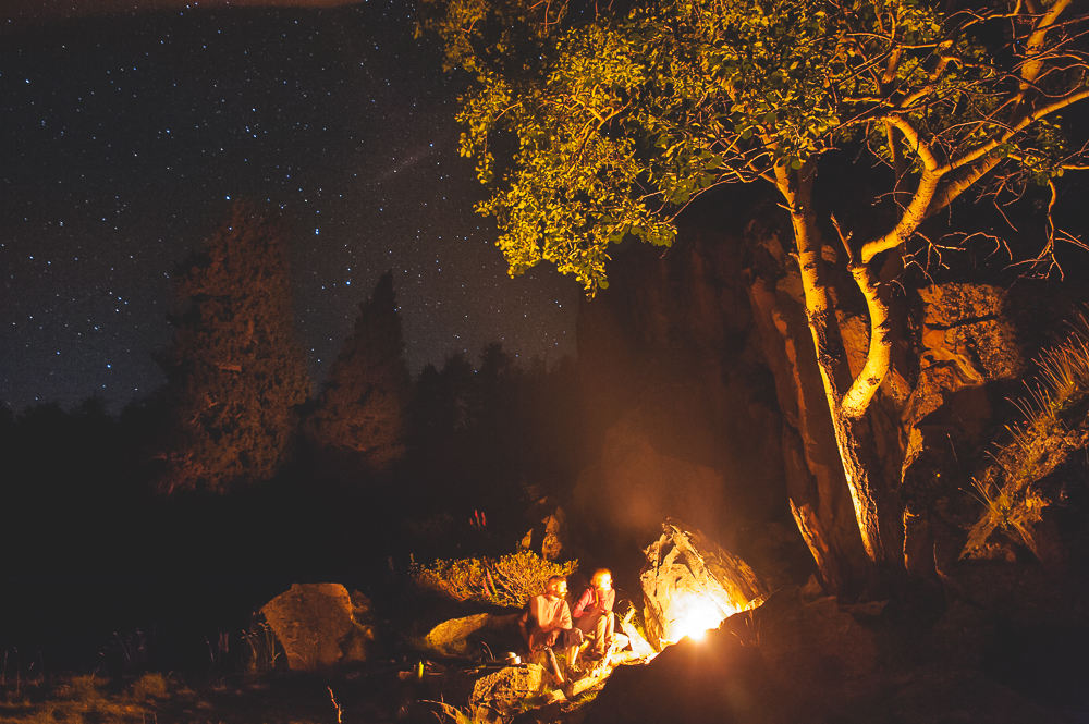 People beside a small fire beneath a warmly lit tree and a dark starry sky