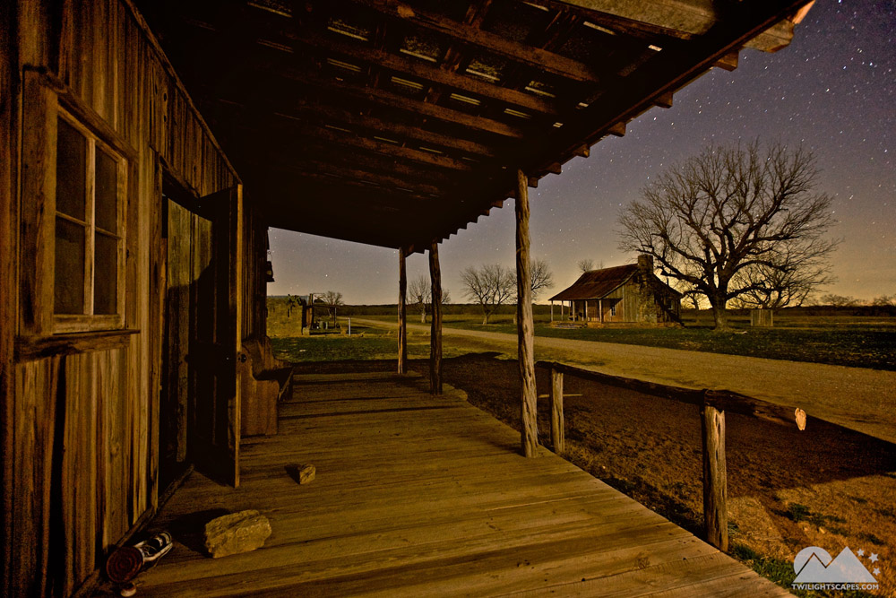 Wooden porch with roof beams and railing facing a lone tree and open land at night