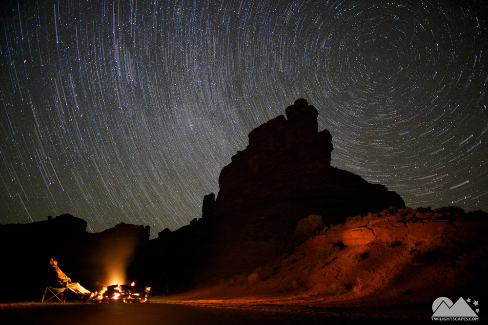 Curved star trails above a dark rock formation with warm foreground light