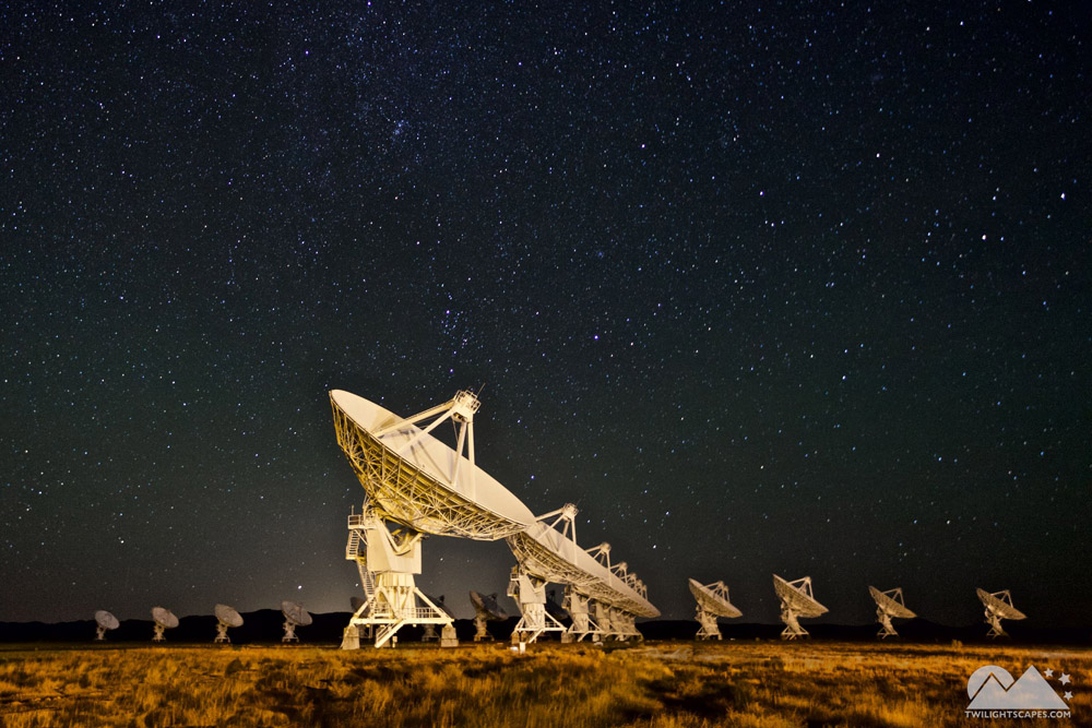 Illuminated radio dishes arranged across a low horizon beneath a wide starry sky