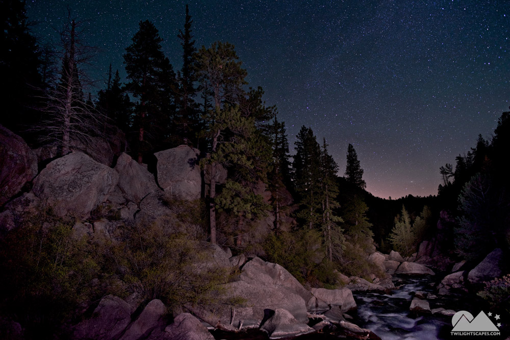 Boulders and trees beside a stream under a darkening blue sky
