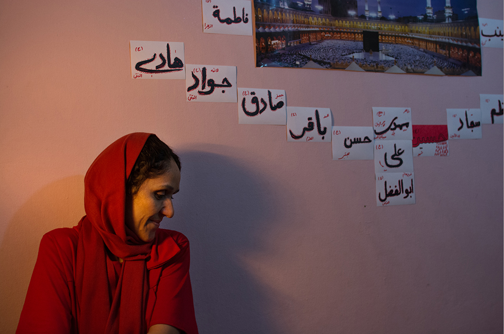 Woman in a red hijab by a wall with Arabic names - The Unblinking Eyes by Nensi Bogdani