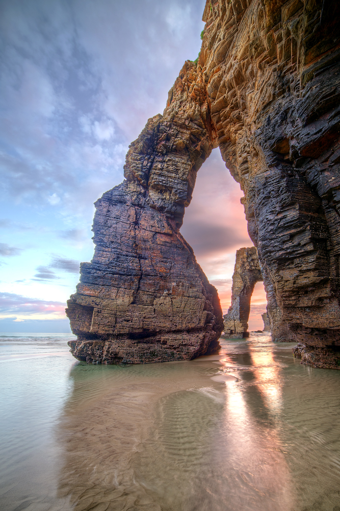 Natural rock arch on As Catedrais beach in Galicia at dusk with wet sand reflections