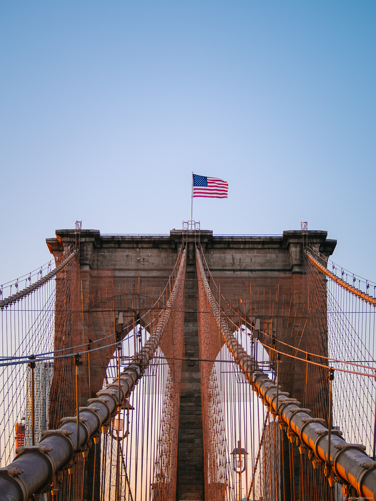 Upward view along suspension bridge cables toward a stone tower and an American flag