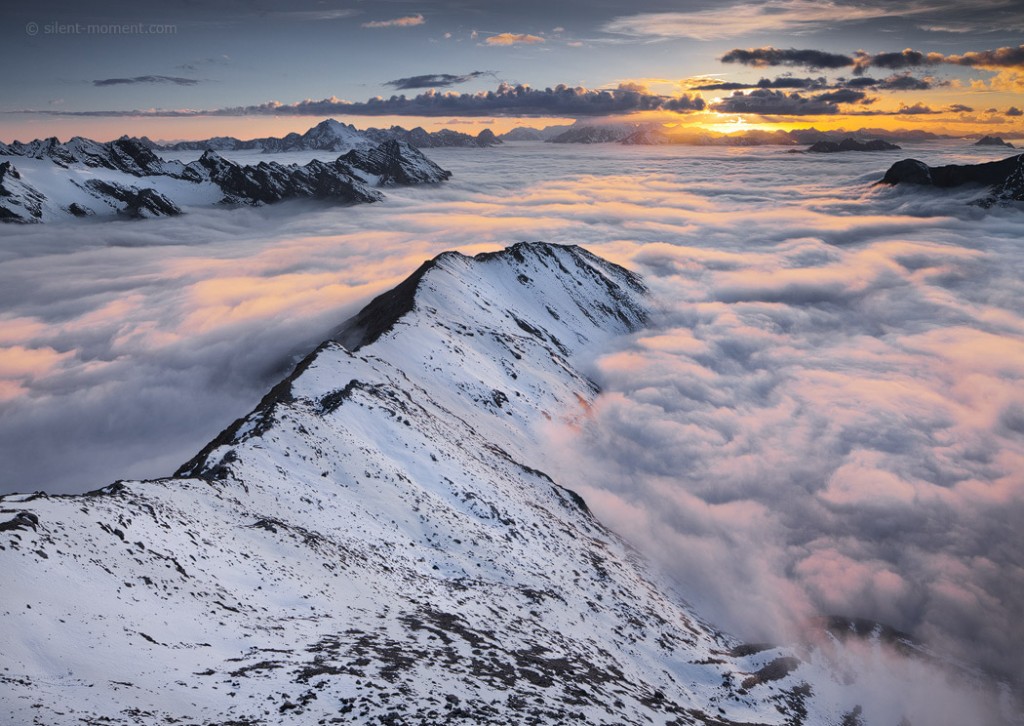 Snow-covered mountain ridge above a broad cloud layer with warm horizon light, viewed from Monte Scorluzzo in Italy