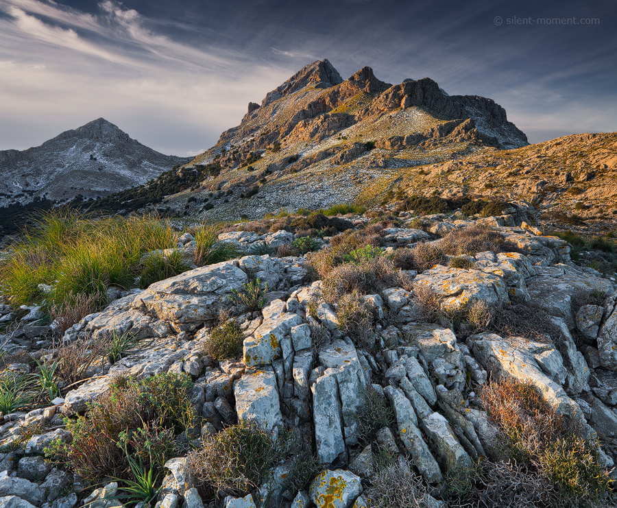 Rocky mountain foreground with low vegetation leading toward a sunlit crag beneath a blue-gray sky