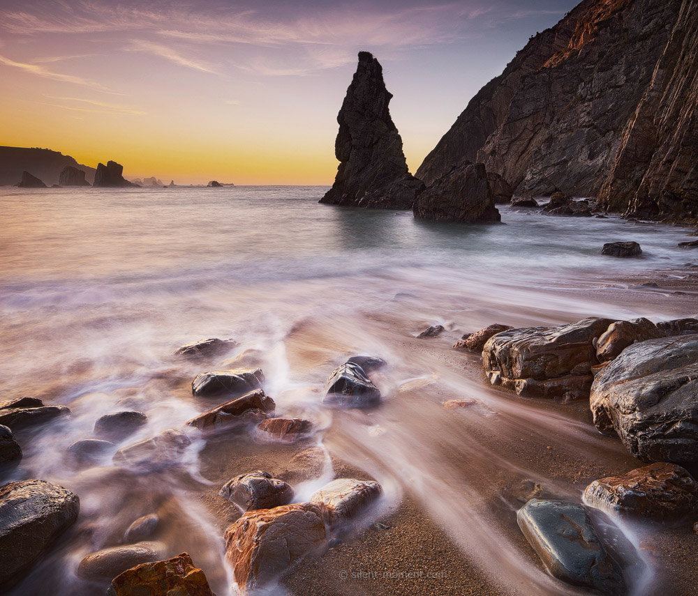 Rocky foreground and a tall coastal sea stack beneath a warm horizon at Playa del Silencio, Asturias, Spain