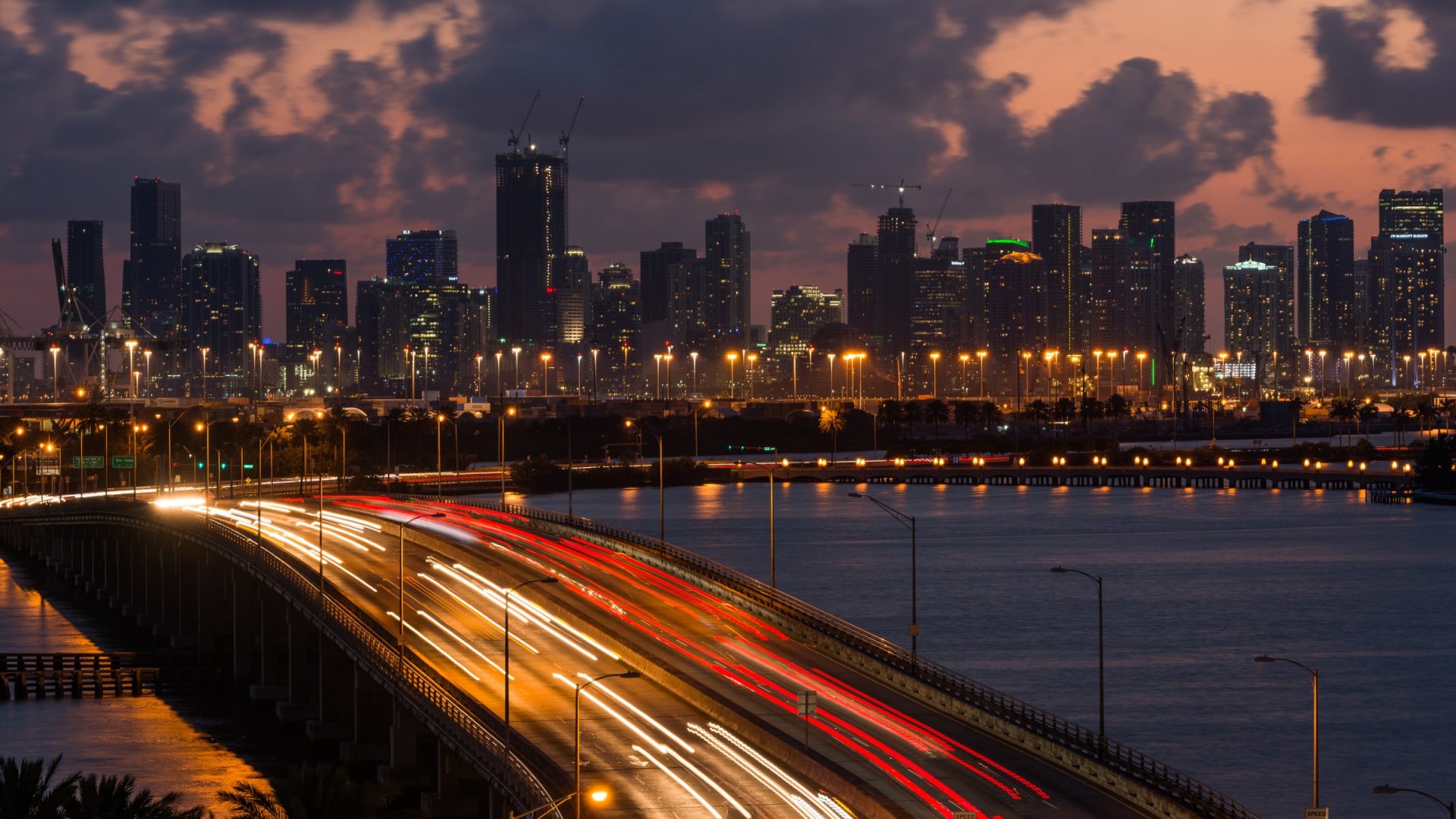 Miami causeway light trails leading toward the downtown skyline beneath pink dusk clouds