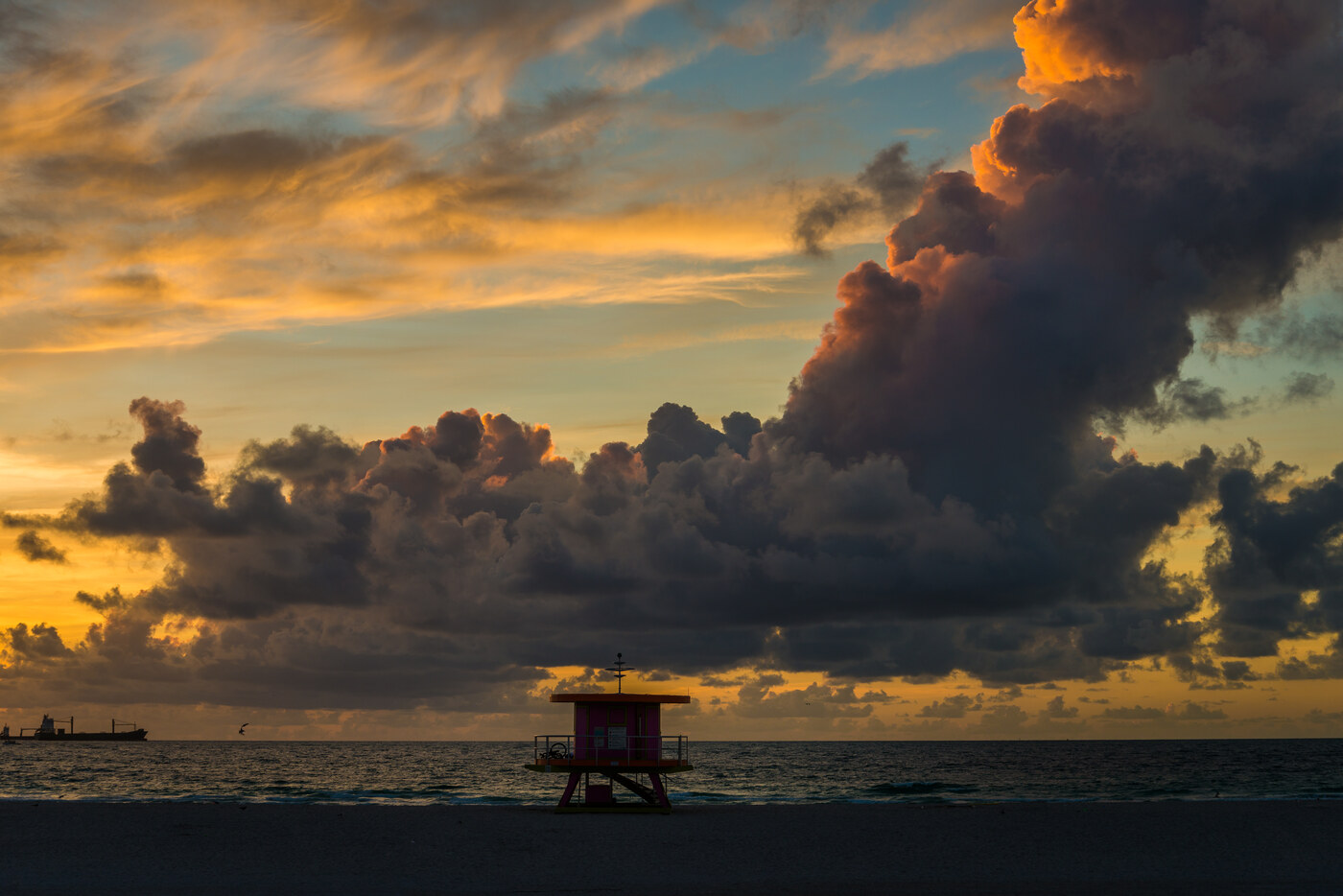 A dark storm cloud lit orange at its edges above a calm sea at sunrise, with a small beach hut on the sand