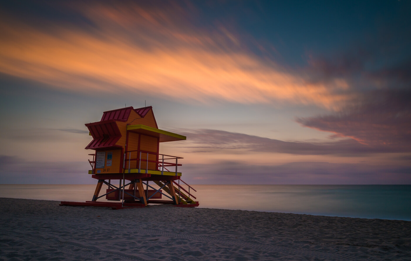 A shaded beach hut with a red roof on sand under orange clouds and a purple sky
