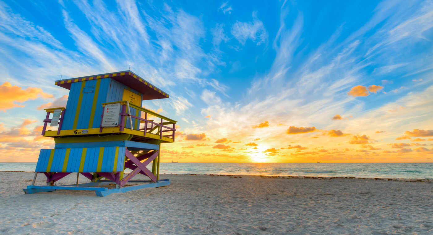 A yellow and blue striped beach hut on sand under a blue sky with streaky white clouds and a low sun on the horizon