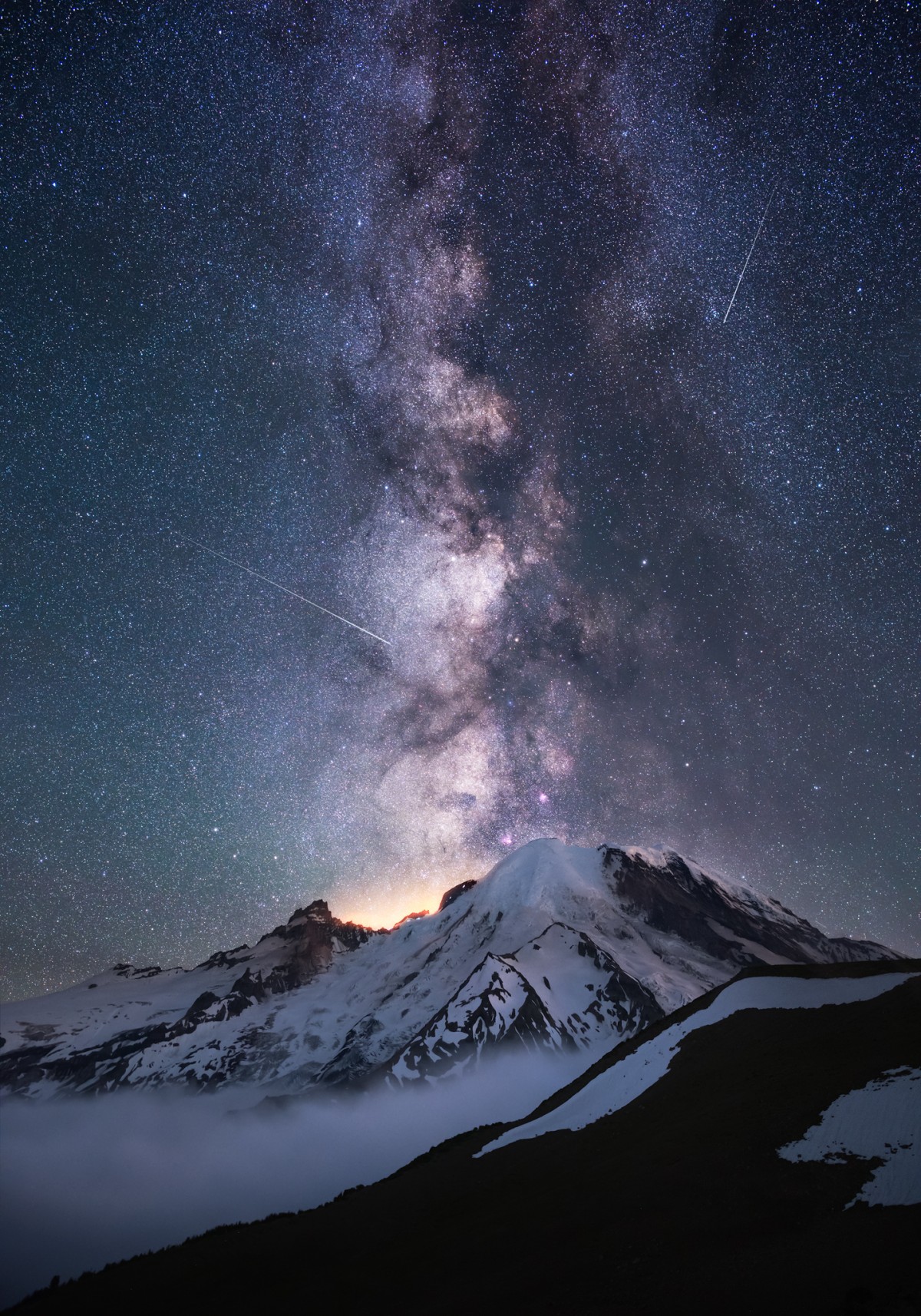 Snow covered mountain below a tall dense star field with bright streaks