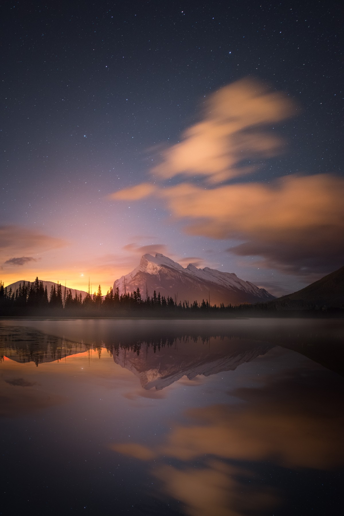 Distant mountain and conifers reflected in still water below warm clouds and a starry sky