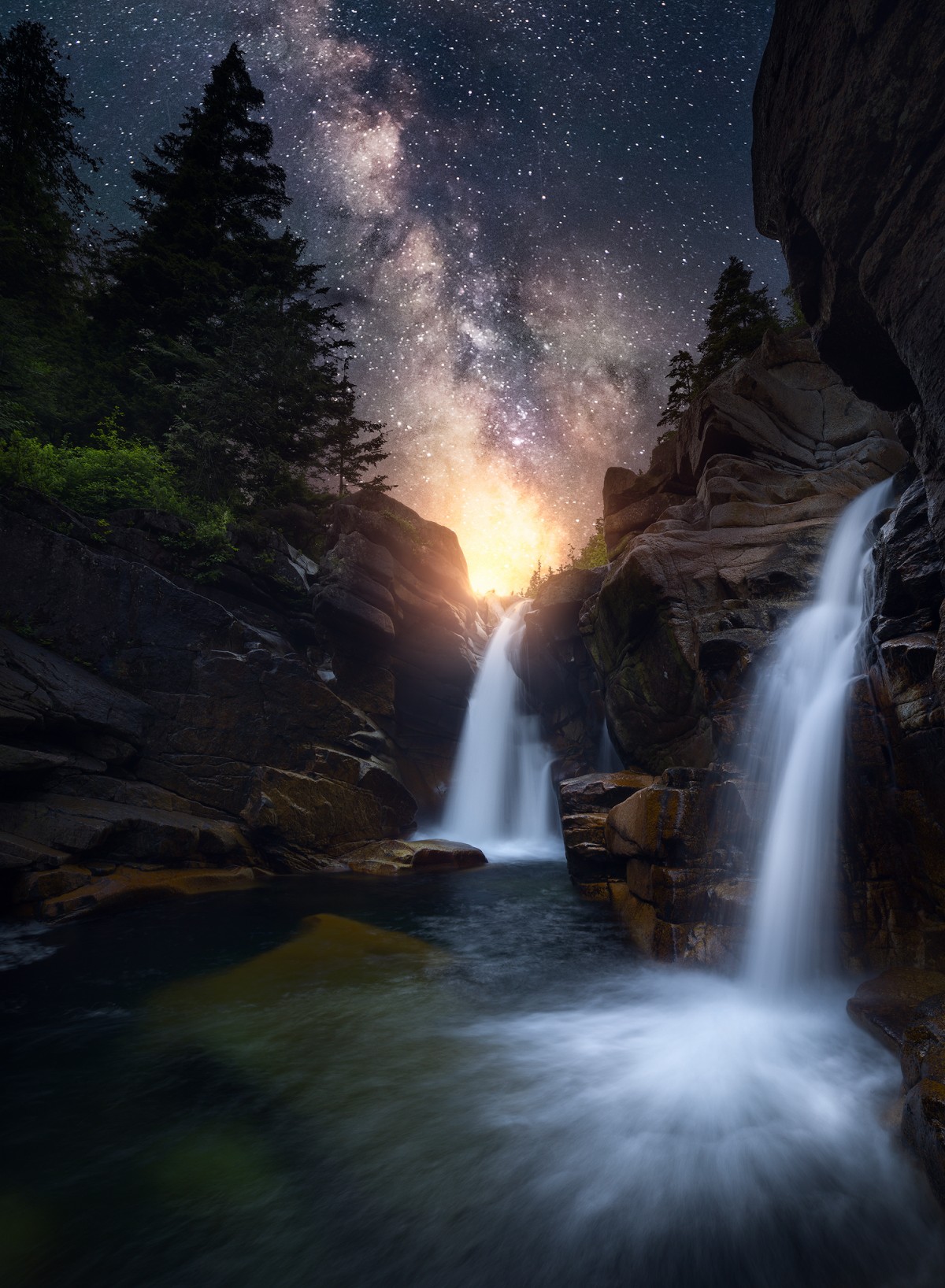 Two waterfalls flowing between dark rocks and conifers below a star filled sky