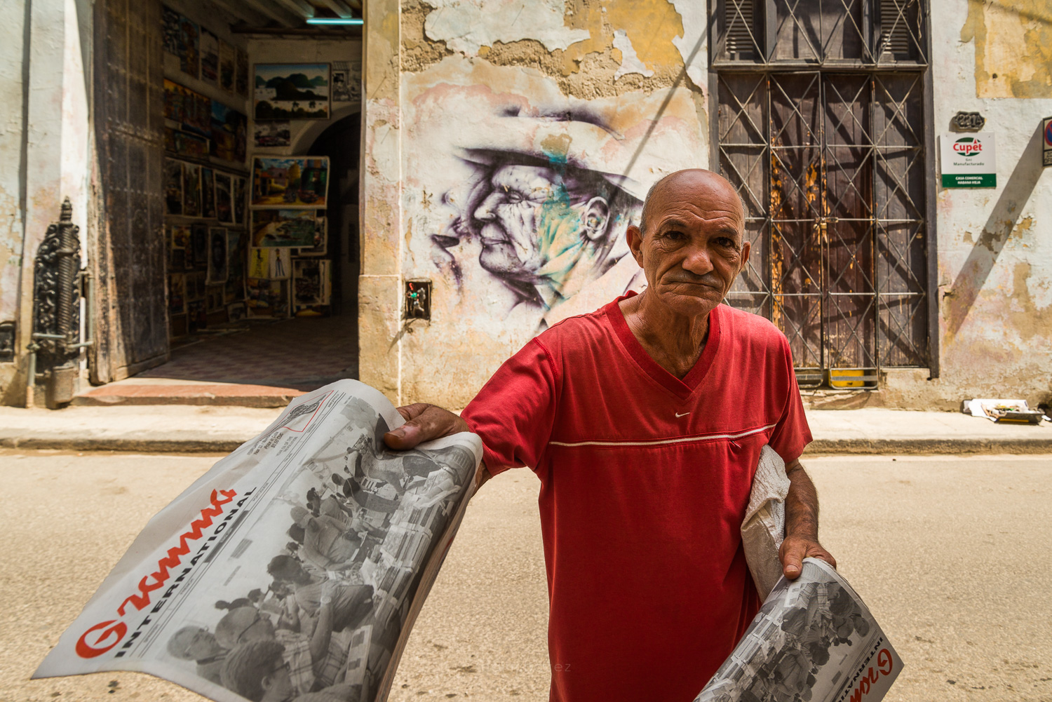Man in a red shirt holding newspapers in front of a weathered wall and face mural