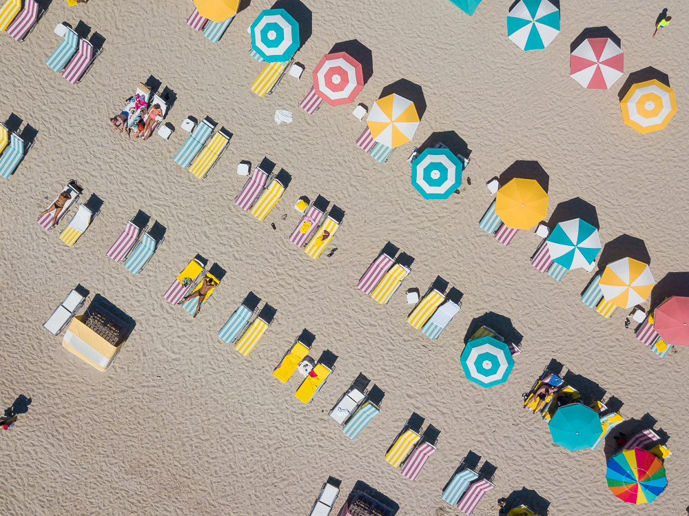 Top-down view of rows of colorful beach umbrellas and striped loungers on sand, with people lying on some of the loungers