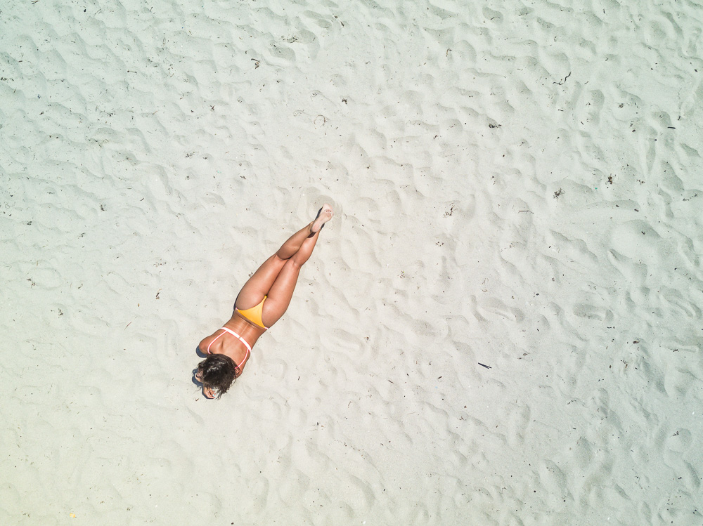 A person in a bikini lying on pale sand seen from directly above, with ripples in the sand around them