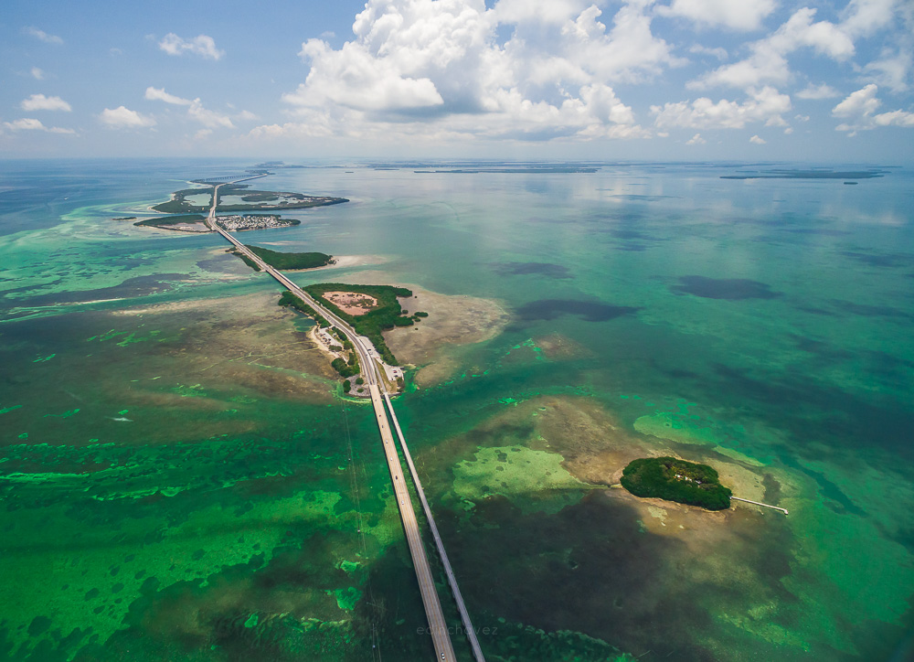 Aerial view of a long highway crossing shallow green and turquoise water with small islands and puffy clouds above