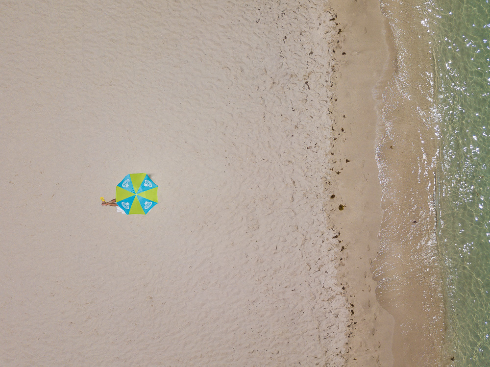 Aerial view of a person lying beside a green and blue umbrella on a sandy beach near the water