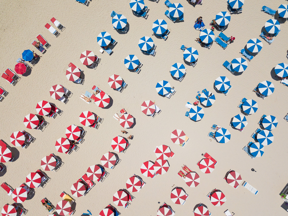 Aerial view of red and blue umbrellas with lounge chairs scattered across a sandy beach