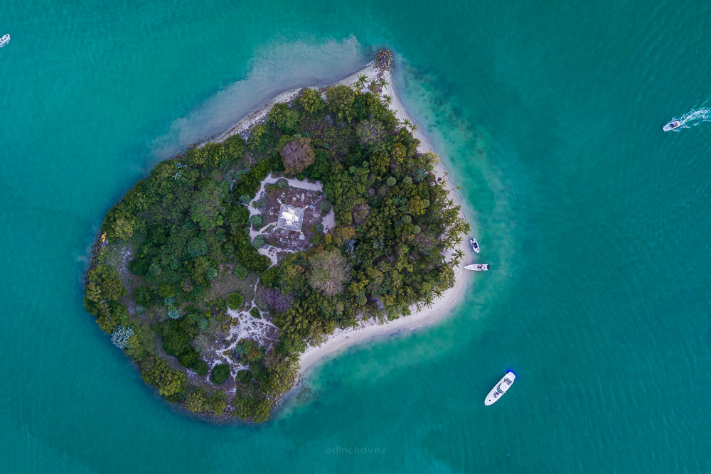 Aerial view of a small tree-covered island surrounded by turquoise water with boats anchored off its beach