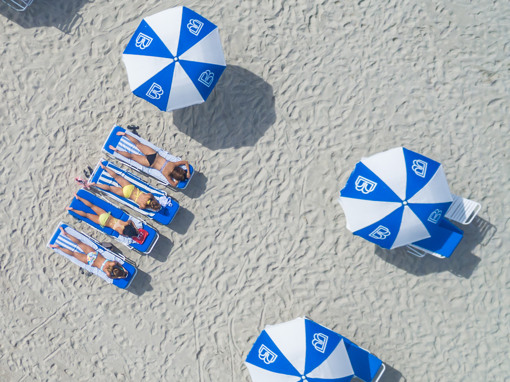 Aerial view of women sunbathing on lounge chairs beside blue and white umbrellas on a sandy beach