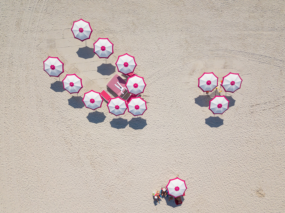 Aerial view of white umbrellas with pink trim clustered around a pink lifeguard stand on a sandy beach