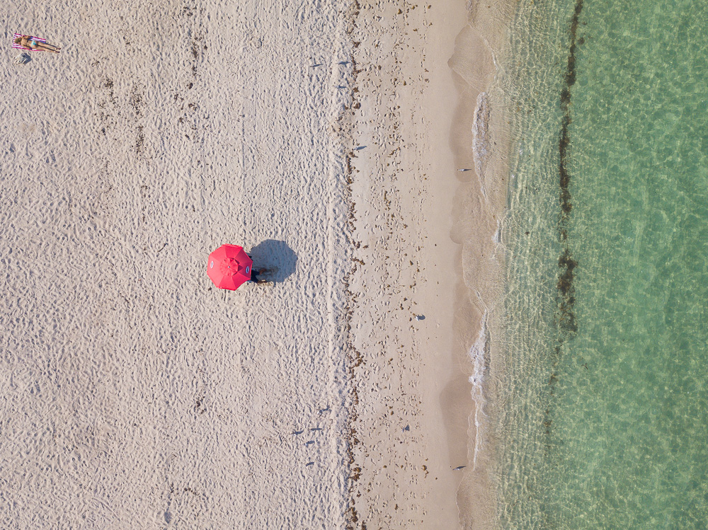 Aerial view of a red umbrella and a sunbather on a sandy beach beside clear green water