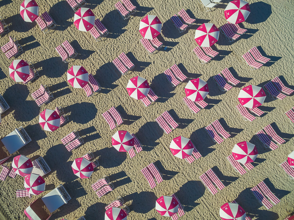 Aerial view of red and white umbrellas and striped lounge chairs casting long shadows on a sandy beach