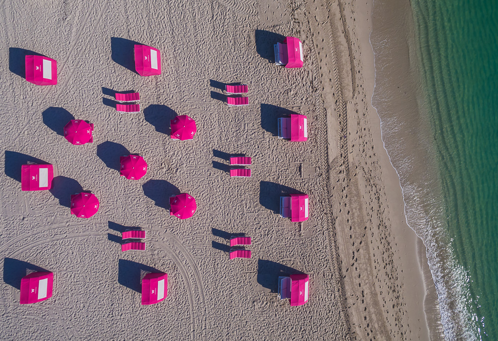 Aerial view of pink cabanas, umbrellas, and lounge chairs arranged on a sandy beach beside green water