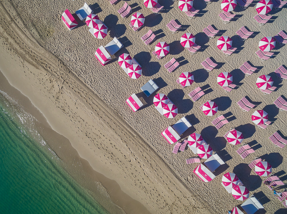 Aerial view of red and white striped umbrellas, lounge chairs, and cabanas along a beach shoreline