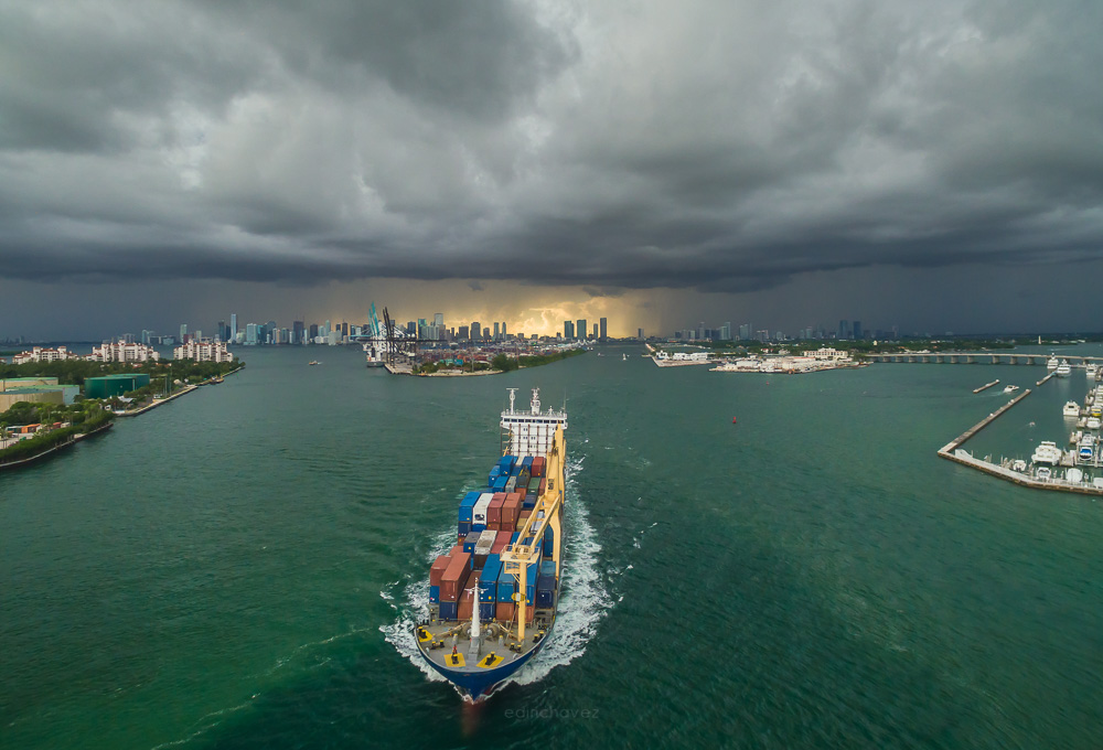 A cargo ship loaded with containers sails toward the camera under dark storm clouds with the Miami skyline in the background