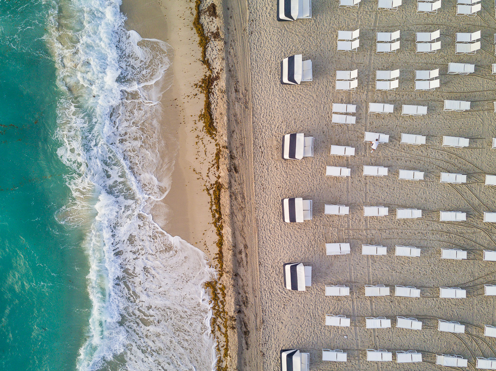 Aerial view of turquoise waves breaking beside rows of white beach chairs and striped tents on sand