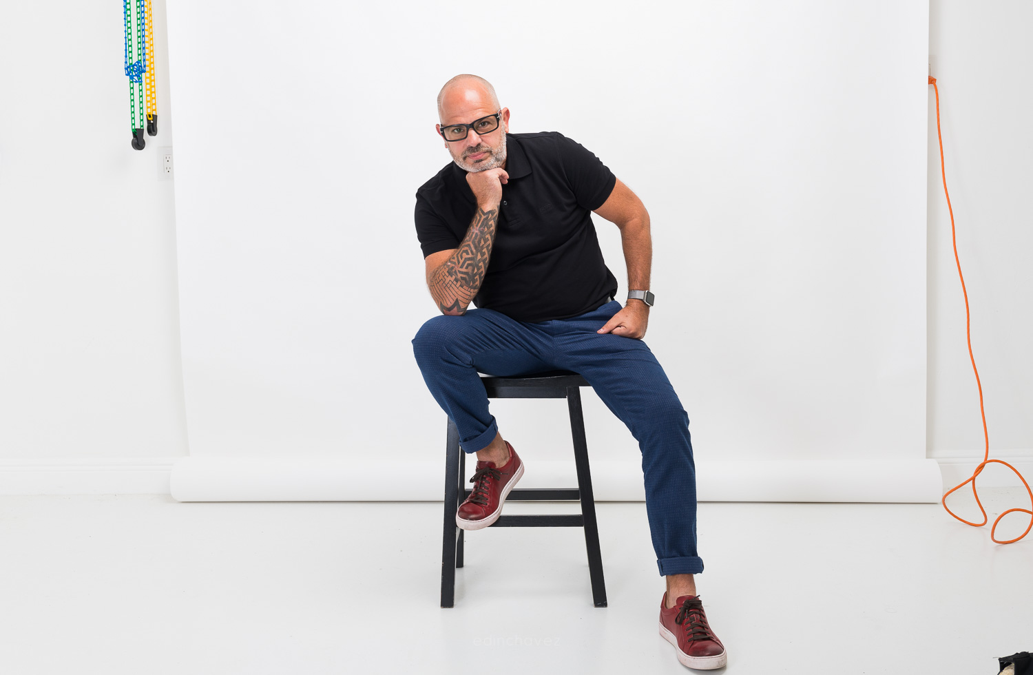 Man with glasses seated on a stool against a white studio backdrop, resting his chin on his hand.
