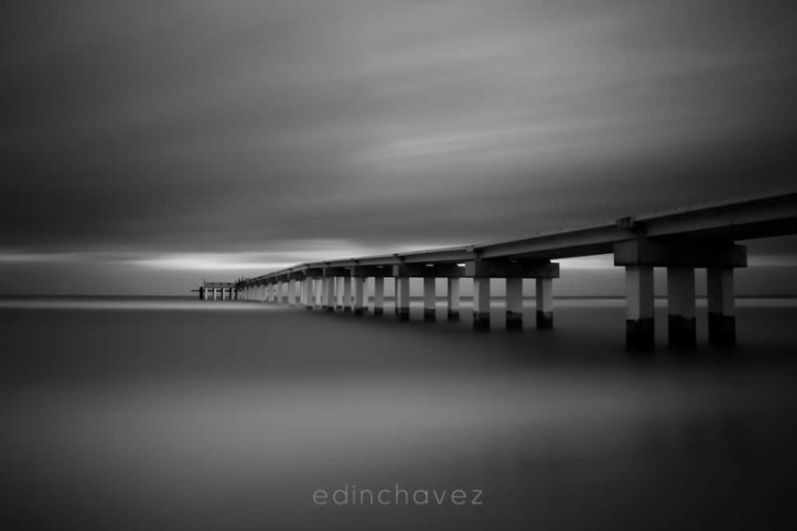 Black and white view of Sunny Isles pier over smooth water under streaked clouds