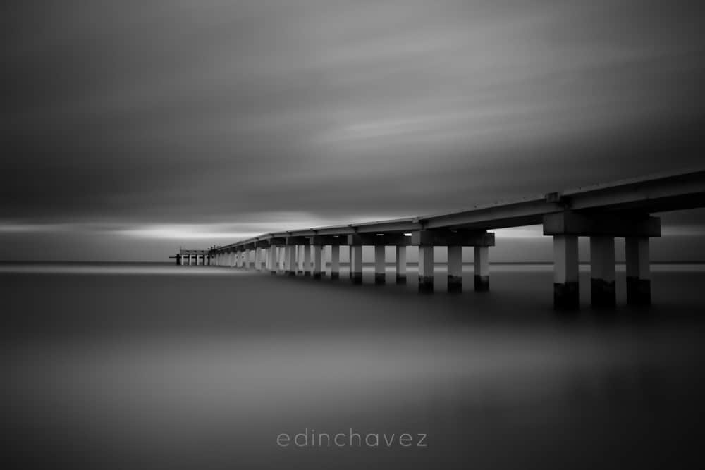 Black and white view of Sunny Isles pier over smooth water under streaked clouds