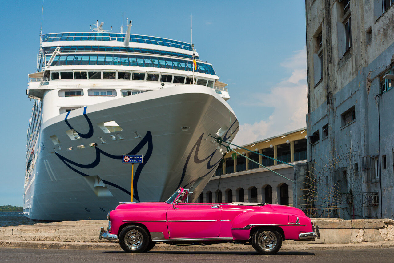 Bright pink vintage convertible beside a docked cruise ship and weathered waterfront buildings in Havana, Cuba