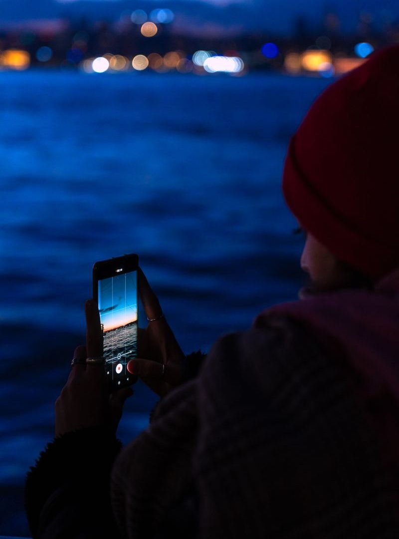 Person holds a phone toward blue twilight water and blurred waterfront lights