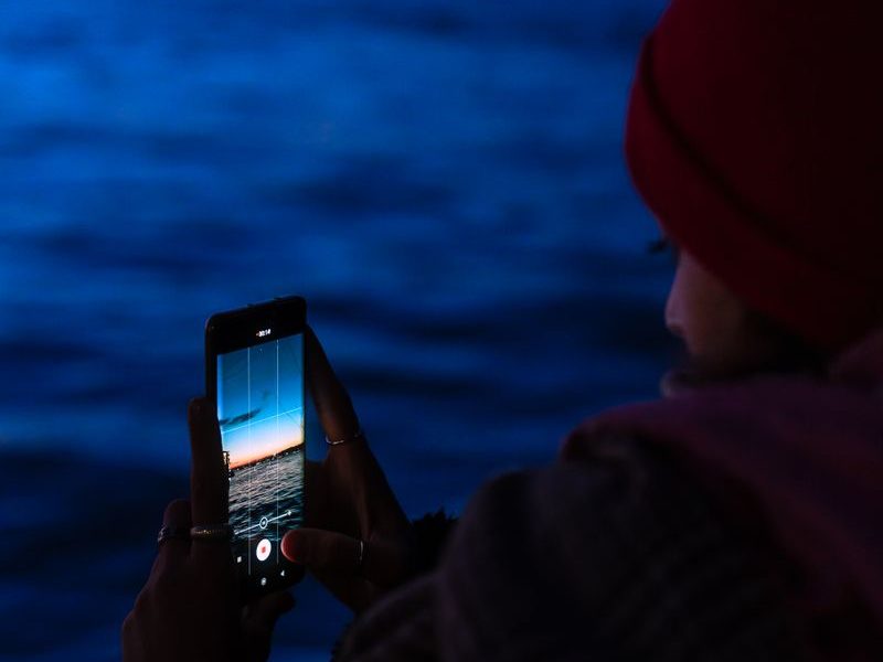 Person holds a phone toward blue twilight water and blurred waterfront lights
