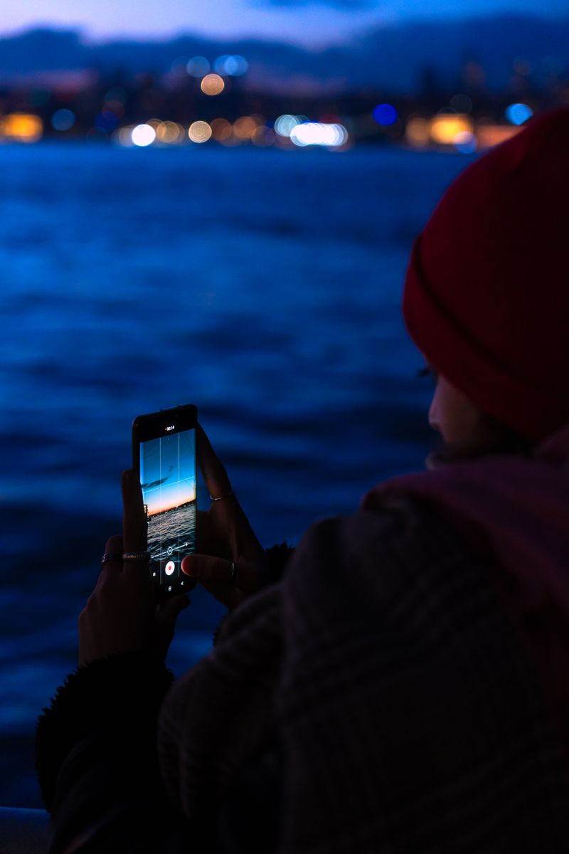 Person holds a phone toward blue twilight water and blurred waterfront lights