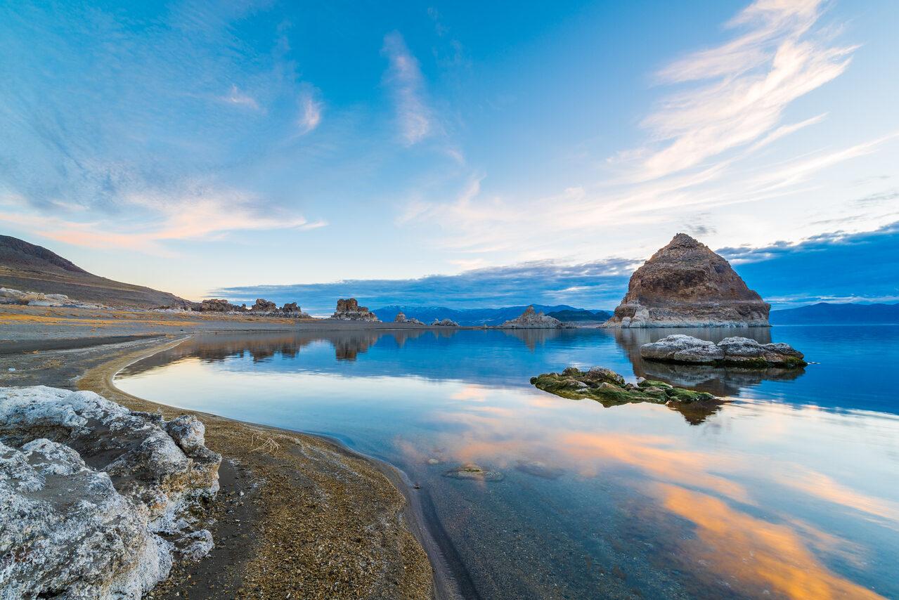 Clouds and blue sky reflected beside the Pyramid Lake shoreline and a large rock formation in Nevada