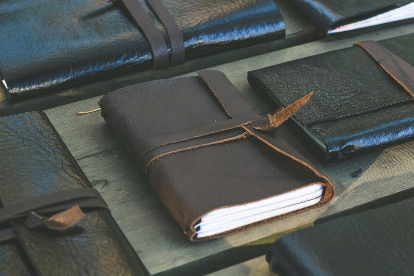 A brown leather-wrapped notebook tied with a strap, its pages visible at the edge, among other dark notebooks on a wooden surface