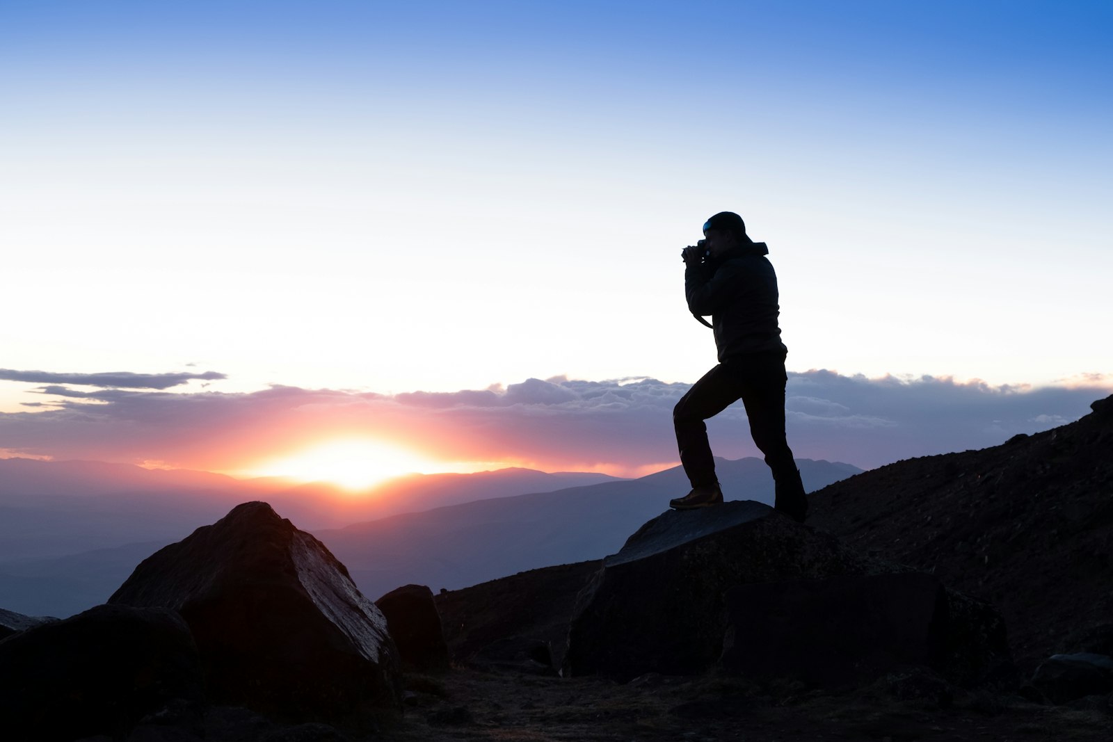 Photographer silhouetted on a rocky mountain at sunset above distant ridges